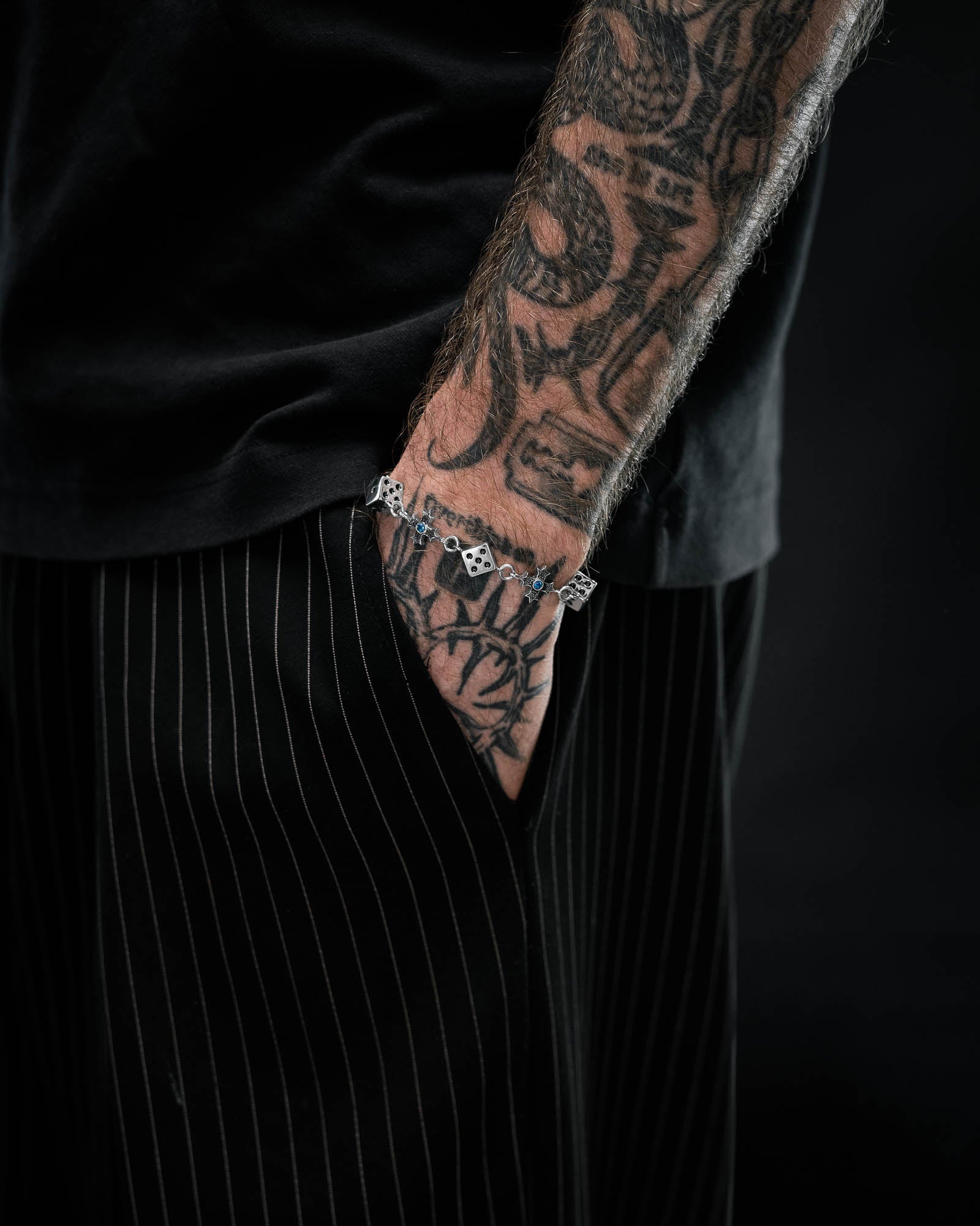 A person with a heavily tattooed arm and hand wears black attire, with their hand in their pocket. On their wrist, the Statement Collective Gambler's Faith Bracelet with stainless steel dice charms is visible.