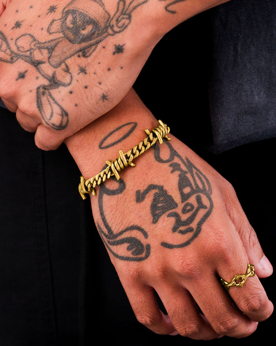 A close-up of tattooed hands wearing the 6mm Barbed Wire Cuban Bracelet (Gold) and a matching gold ring, featuring cartoon tattoos with a halo and hat, set against a black background for a bold jewelry statement.