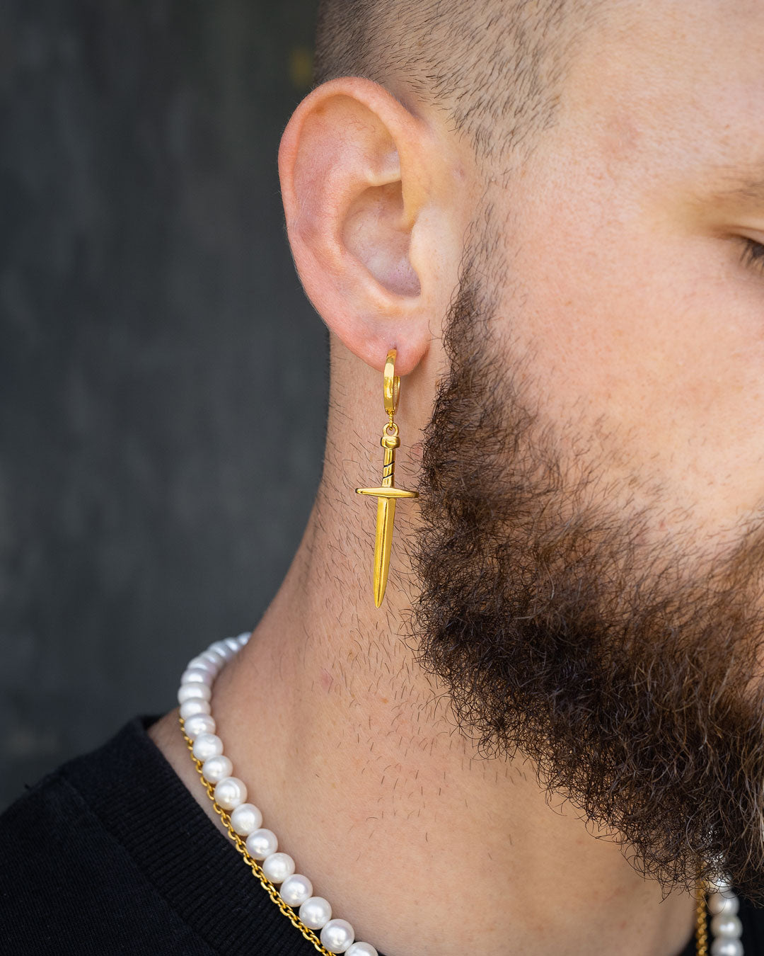 A close-up of a bearded man wearing the Dagger Earring (Gold) and a pearl necklace. Only his ear, part of his cheek, and neck are visible against a dark background.