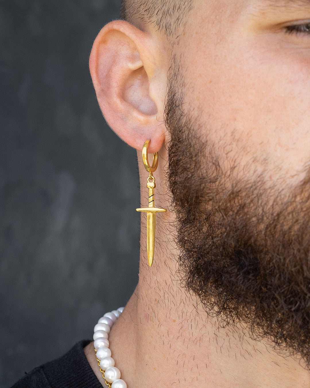 A close-up of a bearded person’s ear wearing the Dagger Earring (Gold); they also have on a necklace with large white beads.