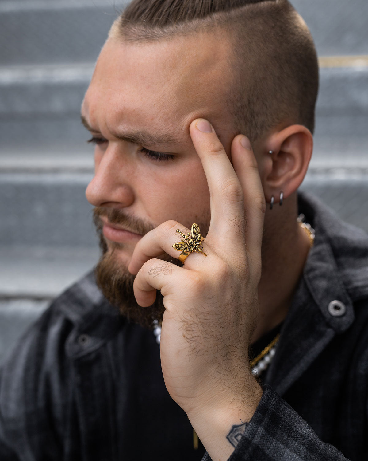 A bearded man with a partially shaved hairstyle sits on stairs, touching his temple with tattooed fingers adorned with gothic jewelry, including chunky earrings and the gold Death's Head Moth Ring, plus a chain necklace and plaid jacket.