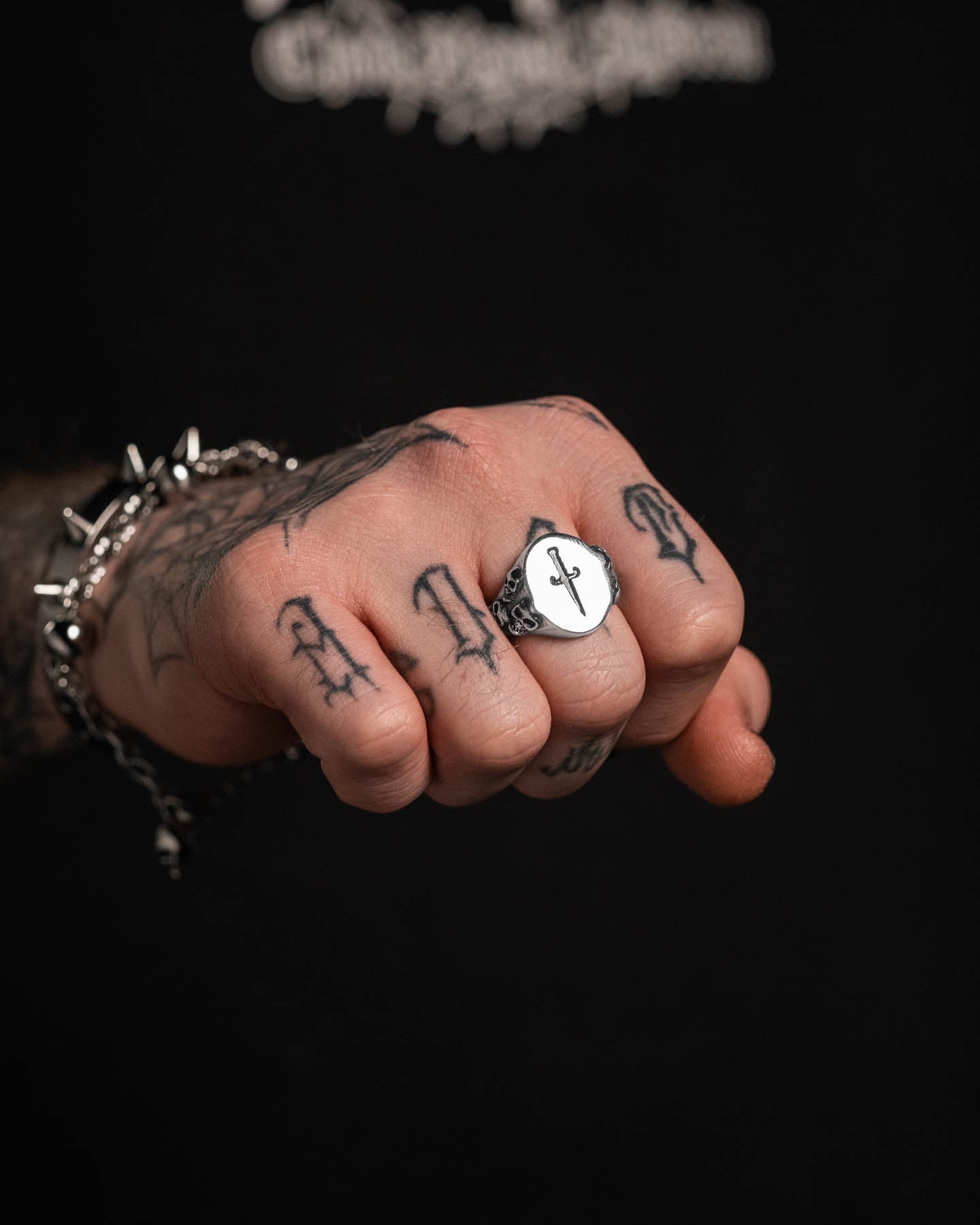 A tattooed hand showcases the Graveyard Signet Ring, a thick stainless steel piece engraved with an anchor, paired with a spiked chain bracelet against a black background.