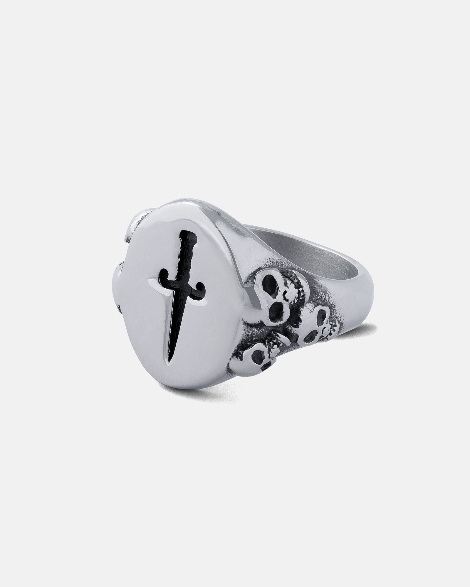 The Graveyard Signet Ring is a silver ring with a round face engraved with a black dagger and a cluster of small skulls on one side, capturing the bold spirit of graveyard jewellery. Shown on a plain, light background.