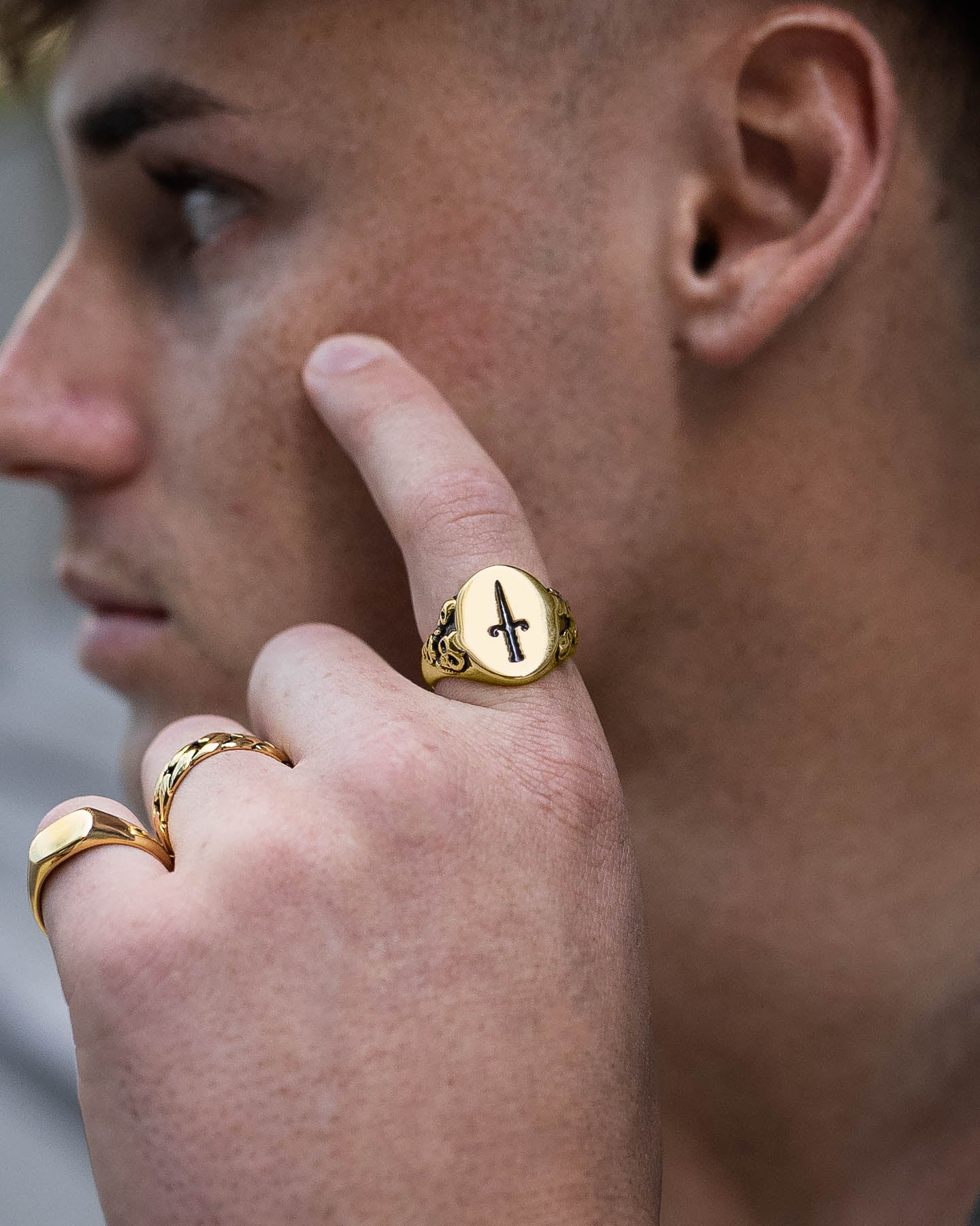 A close-up of a person's face and hand highlights Gothic Jewelry—gold rings, featuring an 18k Gold Skull Ring and the Graveyard Signet Ring (Gold) with a black cross on the middle finger. The person points to their cheek, short hair visible.