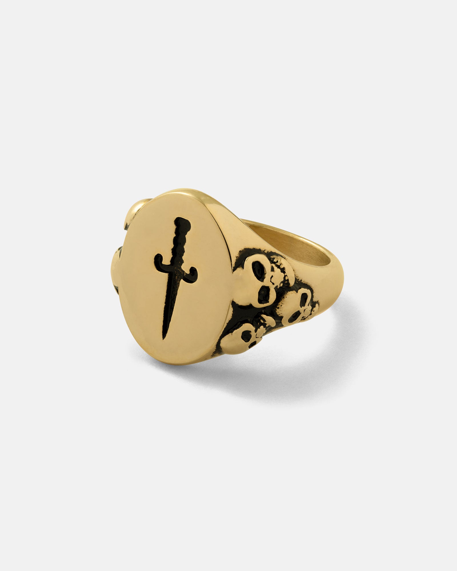 The Graveyard Signet Ring (Gold) is an 18k gold ring with a round face engraved with a vertical dagger and a band adorned with Gothic skulls, shown here on a plain white background.