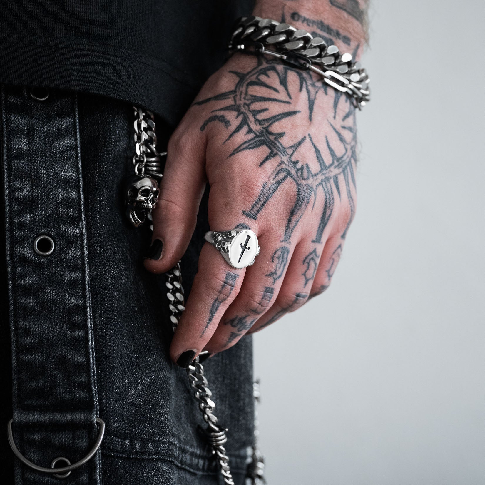 Hand with tattoos wearing a silver signet ring with dagger design through the centre, against a neutral background by statement collective