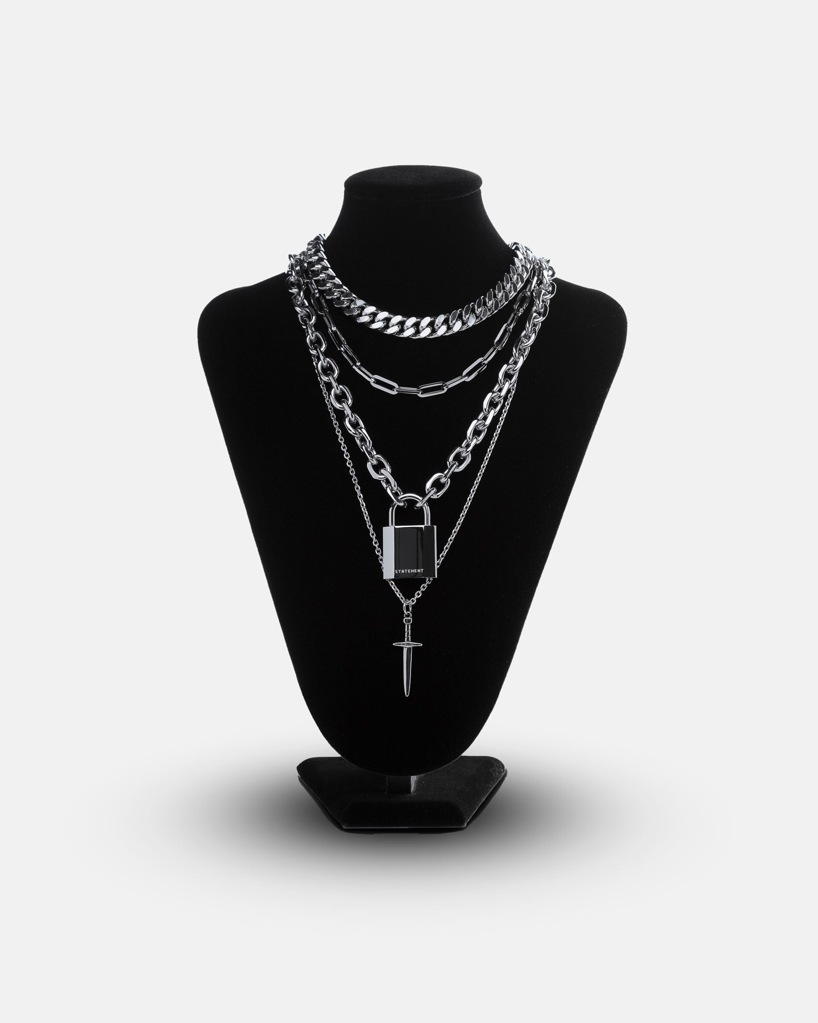 The Handle With Care 2.0 Stack is showcased on a black velvet jewelry bust, featuring layered silver chain link necklaces—chunky curb, elongated links, a rectangular pendant, and a vertical bar—against a white background.