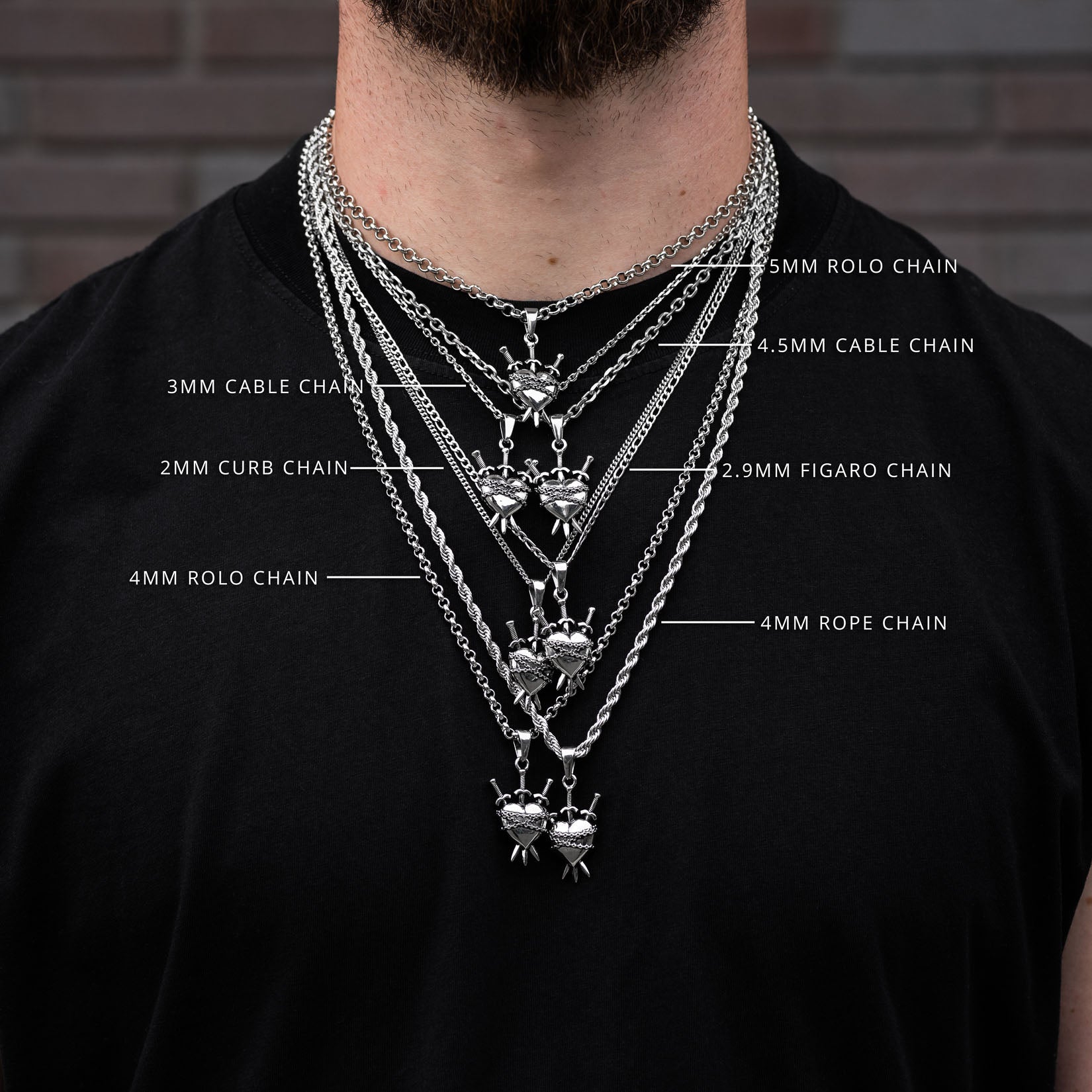 Person wearing multiple silver heart chains with chain type labels on a black shirt against a brick wall.
