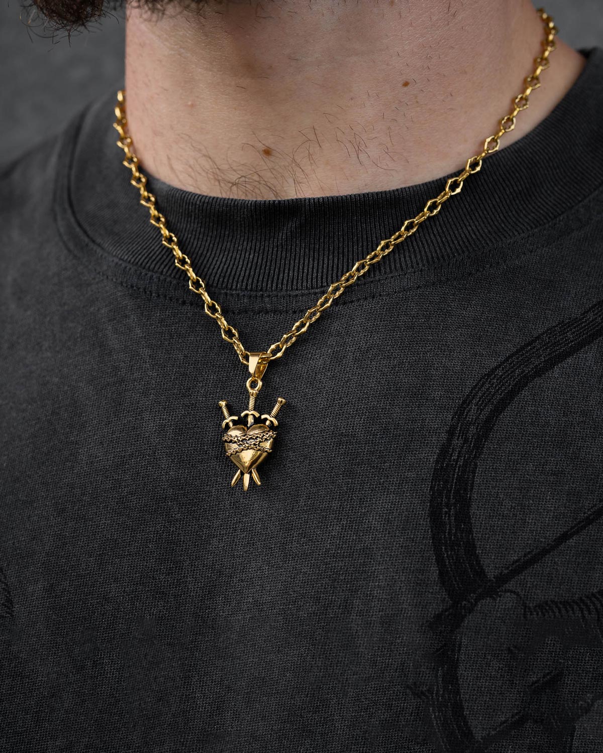 A person wears Statement Collective's "The Cathedral" 6mm Spiked Chain in gold with a custom heart-shaped pendant featuring three swords and a crown. The lower face, neck, and chest are visible, highlighting the bold gothic accessory.