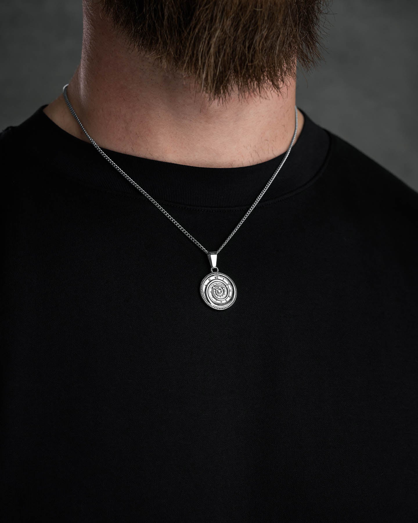 A person wears the Infinite Clock Pendant Necklace—a silver spiral clock design in 316L stainless steel—with a black shirt; only their lower face, beard, neck, and upper chest are visible against a dark background.
