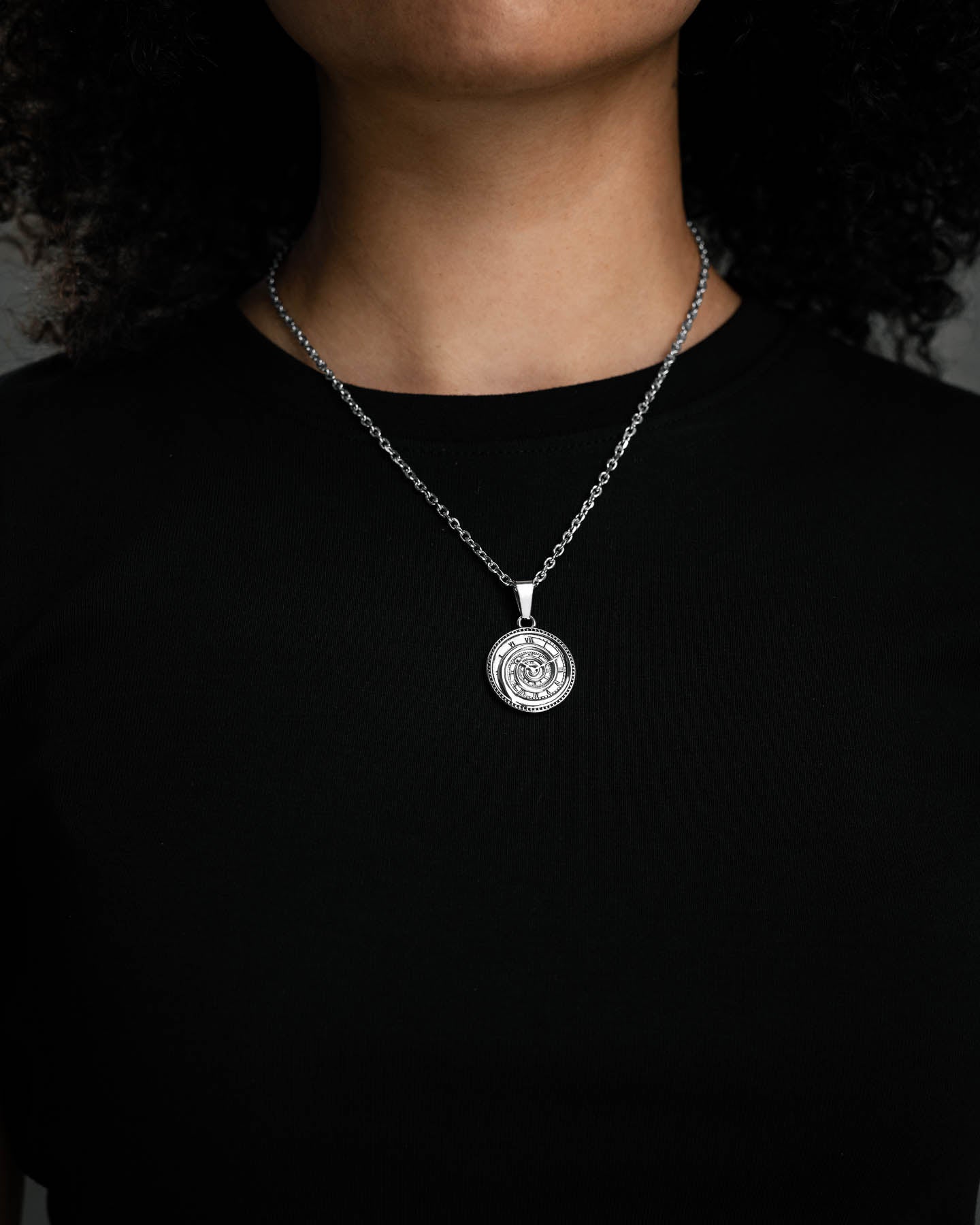A person wearing a black shirt and the Infinite Clock Pendant Necklace—featuring a Dali-inspired spiral clock design crafted from 316L stainless steel—against a dark background. The focus is on the detailed pendant.