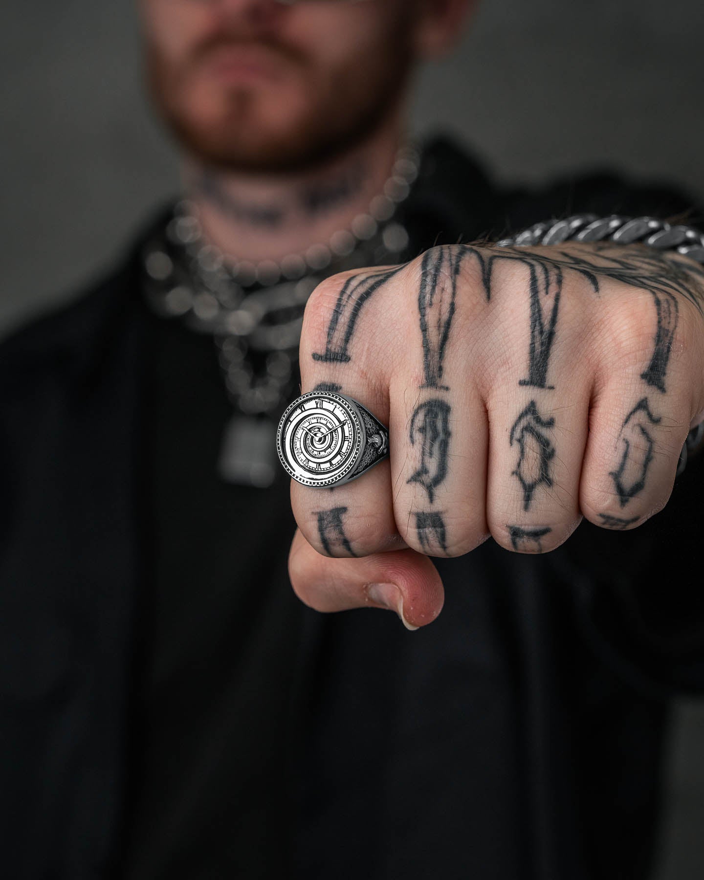 A tattooed hand wearing the Infinite Clock Ring—a large spiral ring with intricate detailing—extends a fist toward the camera. A chain bracelet is also worn, and the background is blurred.