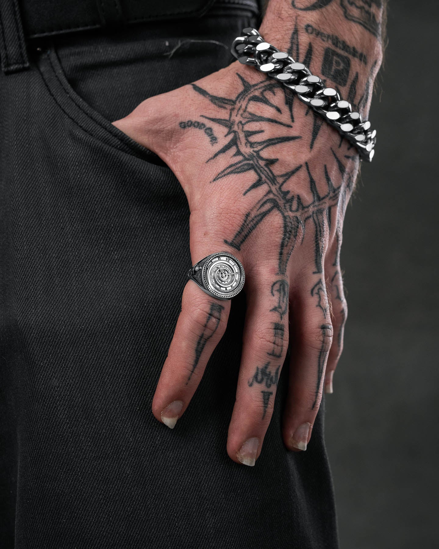 A close-up of a tattooed hand on black jeans, wearing the Infinite Clock Ring and a chunky silver chain bracelet, highlighting the intricate jewelry design.