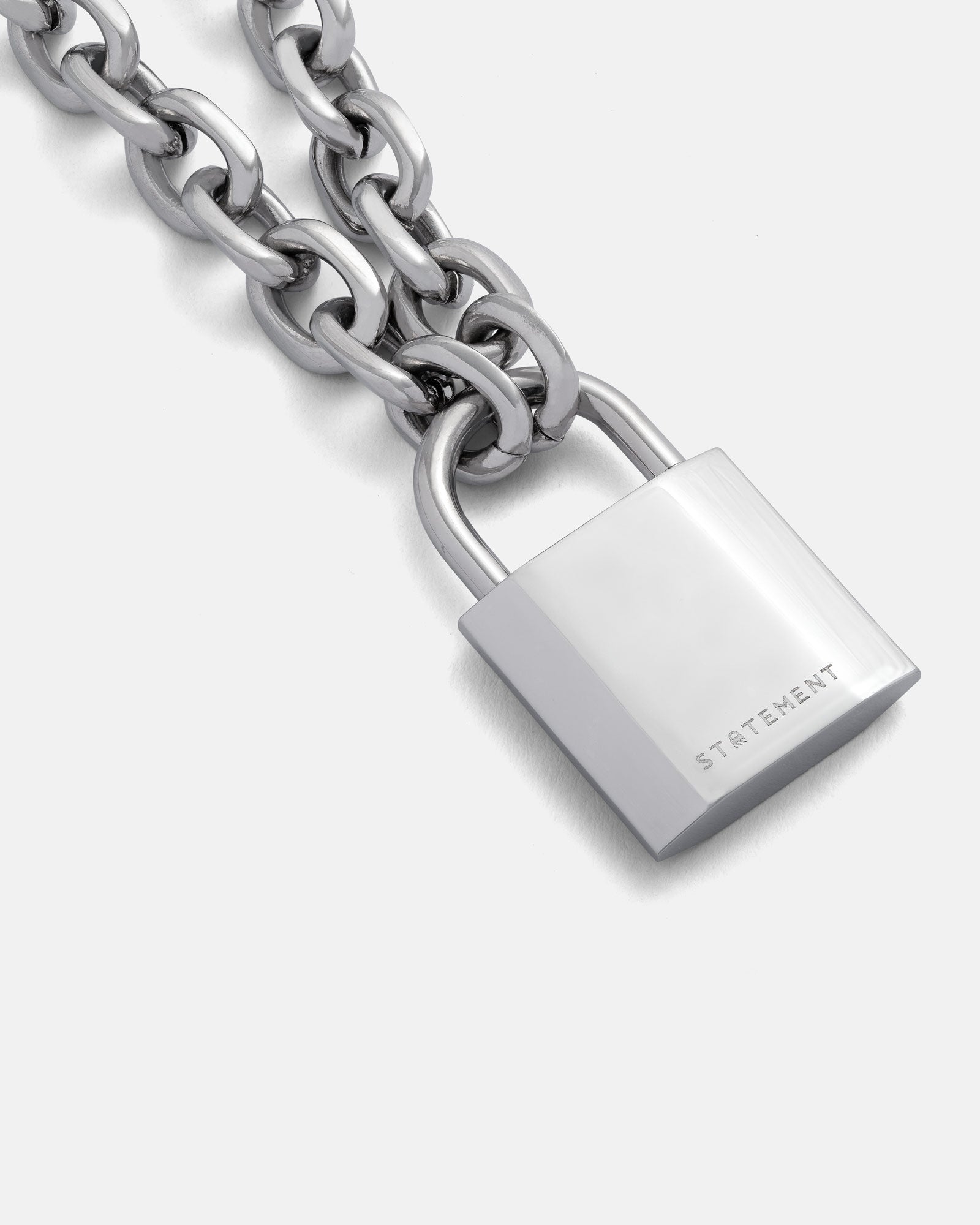 The Statement Lock & Chain 2.0 is a chunky silver necklace with a shiny, rectangular padlock engraved with "STATEMENT," shown against a plain light gray background.