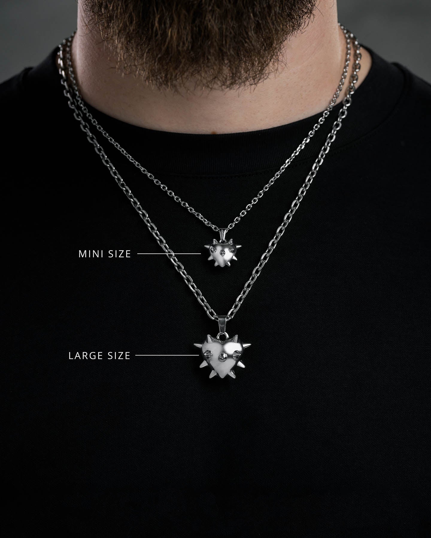 A person wears two "Love Hurts" Spiked Heart Pendants—Mini Size and Large Size—layered over a black shirt, ideal grunge jewelry for fans of the bold, edgy spiked heart style.