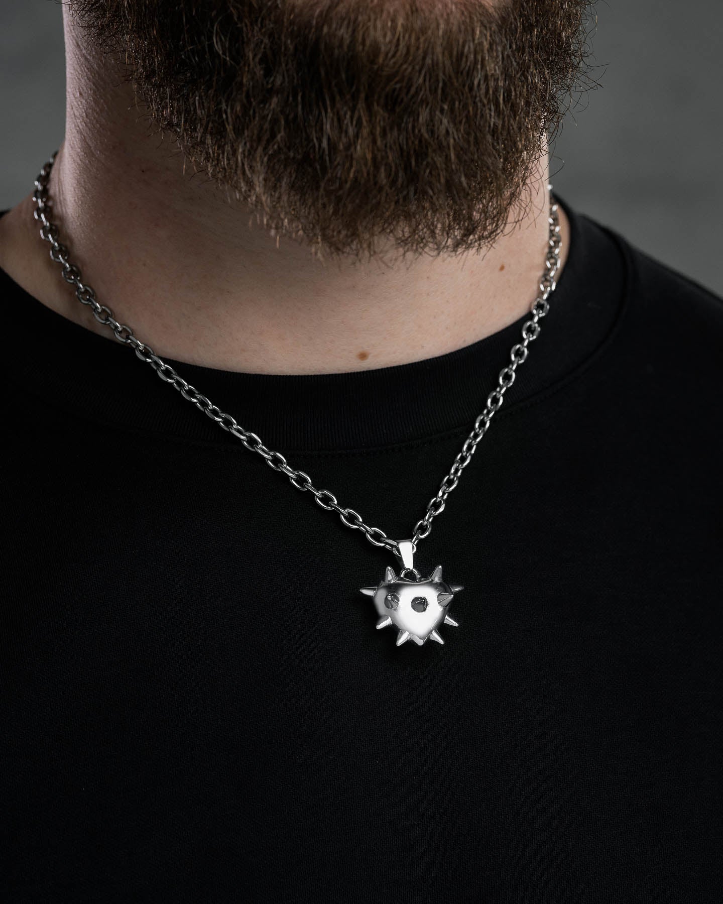 A close-up of a person with a brown beard wearing a black shirt and silver chain necklace featuring the "Love Hurts" Spiked Heart Pendant, blending edgy grunge style with a morning star-inspired design.