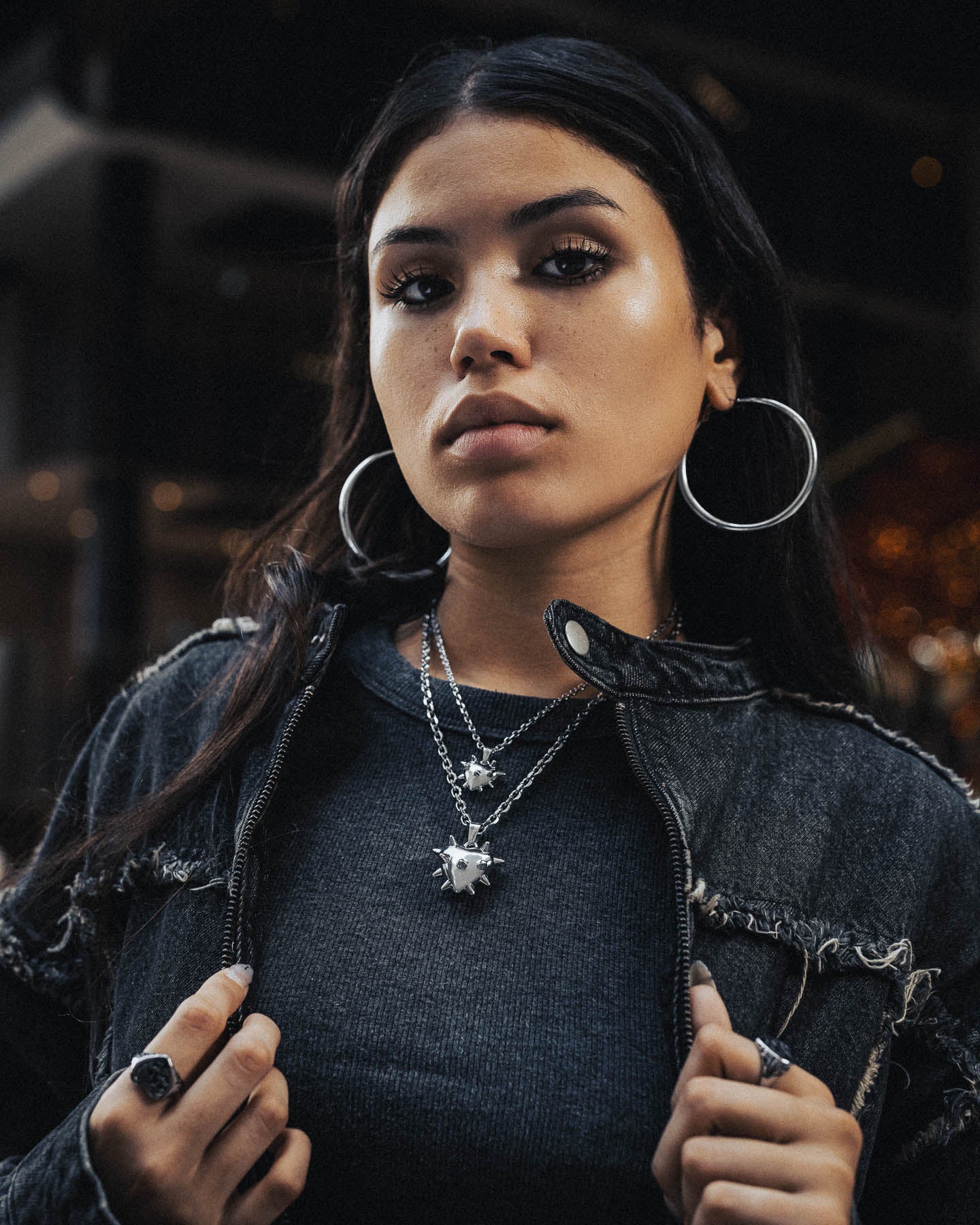 A woman with long dark hair and hoop earrings wears grunge jewelry, including the "Love Hurts" Spiked Heart Pendant, layered silver necklaces, rings, and a distressed black denim jacket as she looks confidently at the camera against a dark blurred background.