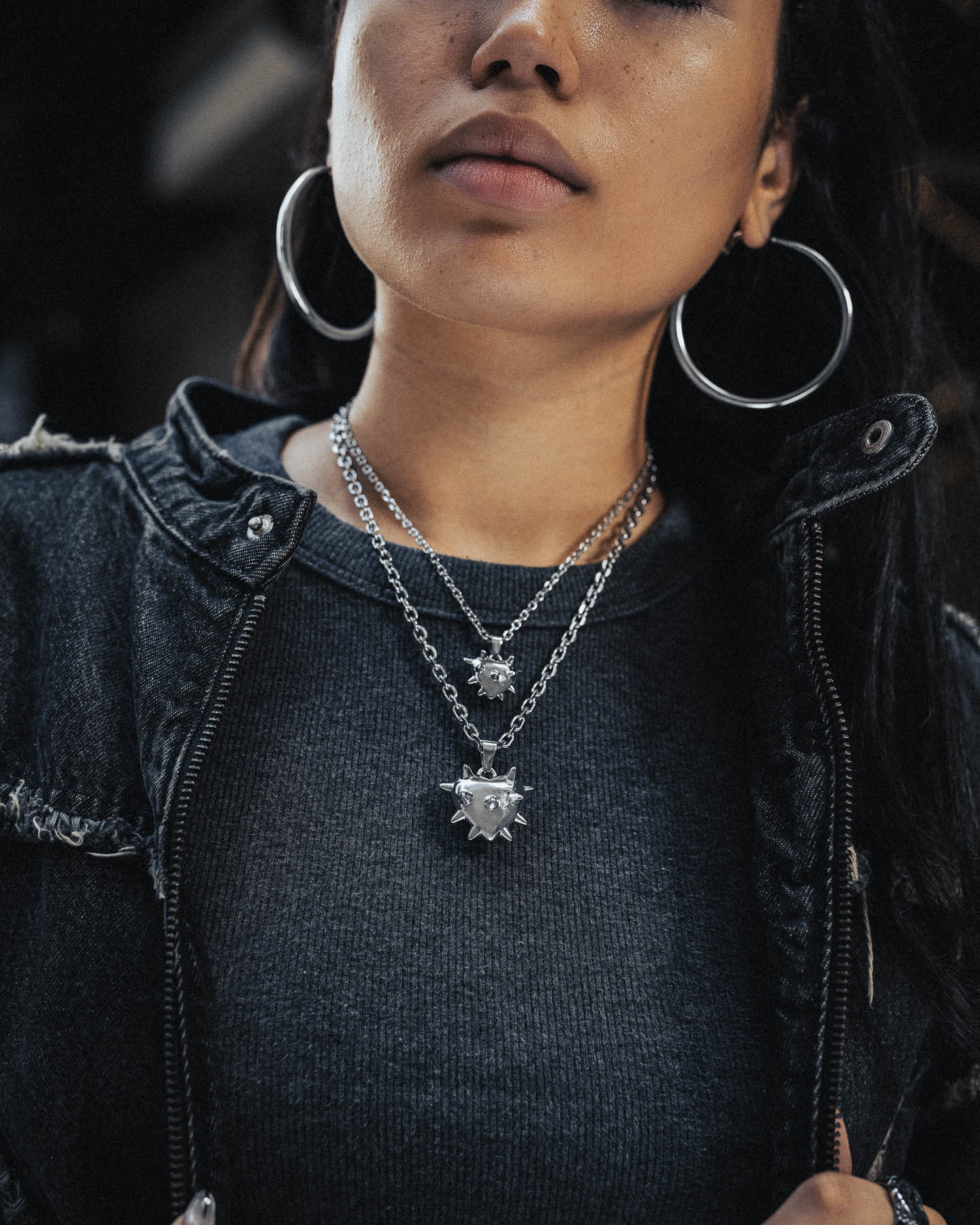 A person wearing large hoop earrings, the "Love Hurts" Spiked Heart Pendant with layered silver chains, a ribbed gray top, and a distressed black denim jacket. The photo is cropped to show only the lower face and upper torso.