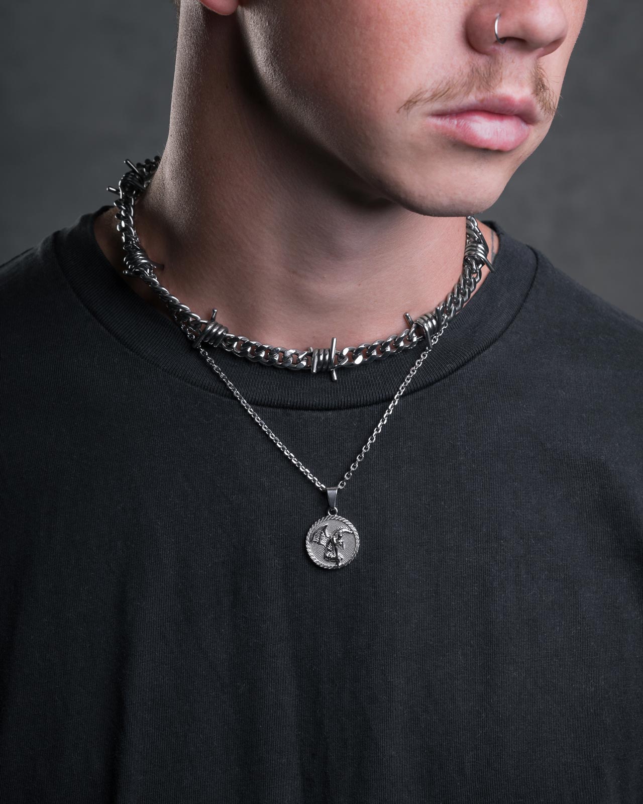 A person wearing a black t-shirt, barbed wire-style chain, and Statement Collective's Fallen Angel Medallion Pendant. The image highlights their nose ring, mustache, and the pendant on their upper chest.
