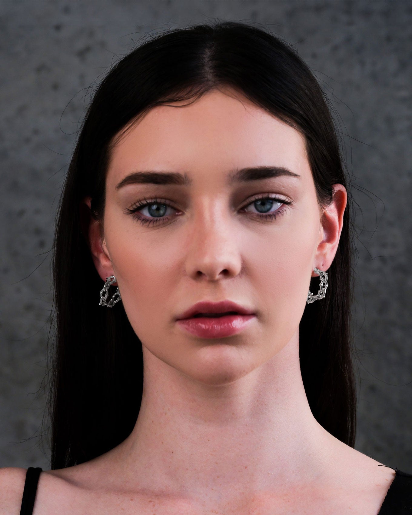 A woman with long dark hair and blue eyes, wearing "The Cathedral" Hoop Earrings and a black strap, faces the camera with a neutral expression against a gray textured background.