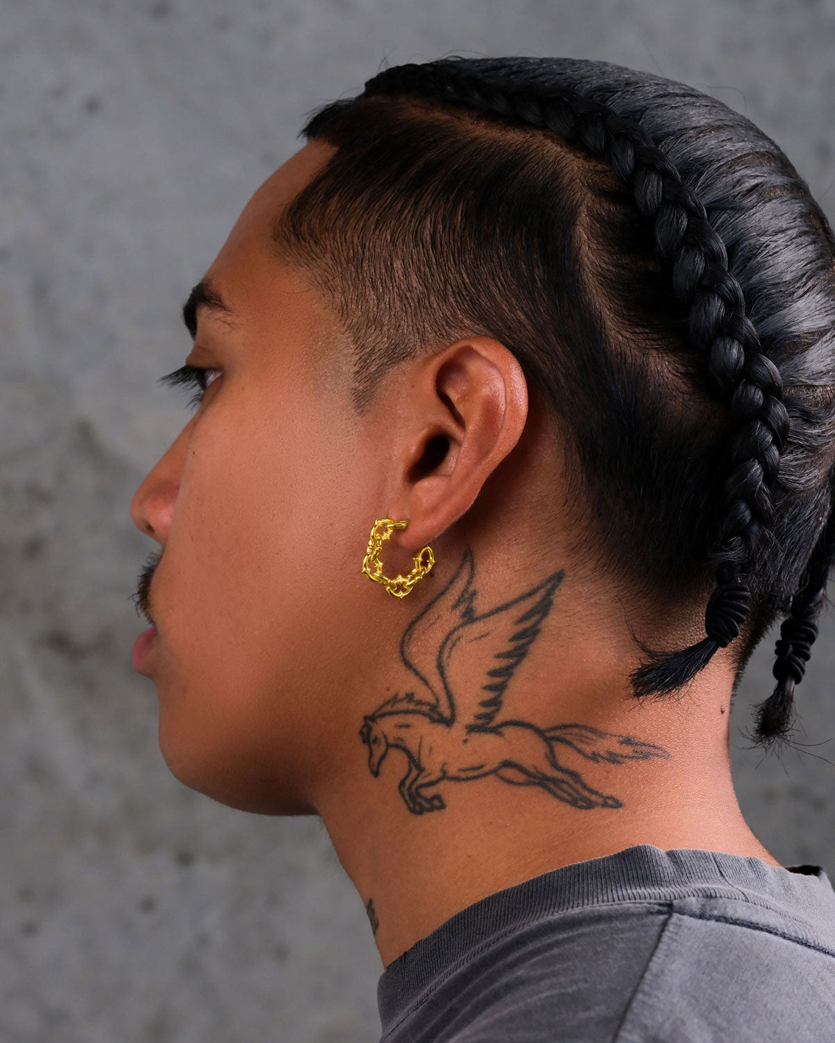 A person with braided hair, a mustache, and a winged horse neck tattoo wears "The Cathedral" Hoop Earrings (Gold) against a plain gray concrete wall.