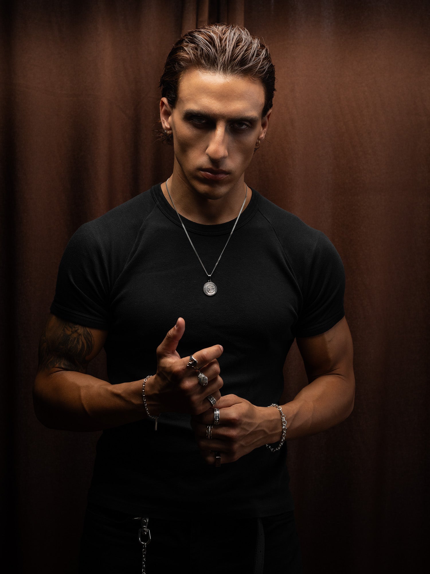 Man wearing a black t-shirt and clock medallion necklace against a brown background