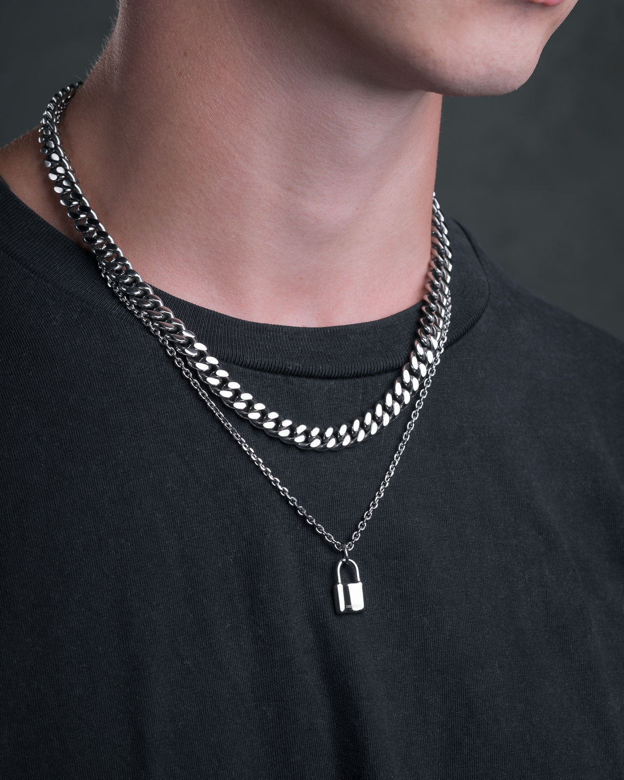 A person wearing a black t-shirt, styled with two silver chain necklaces: a thick curb chain and a 2mm chain featuring the Mini Padlock Pendant.