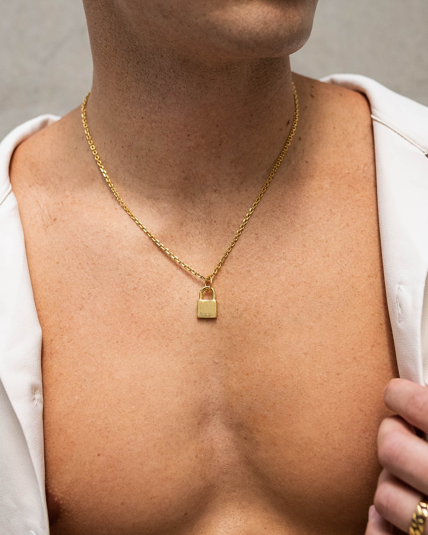 A close-up of a bare chest wearing the Mini Padlock Pendant (Gold), a hypoallergenic 18k gold necklace that shines beneath an open white shirt draped over the shoulders.