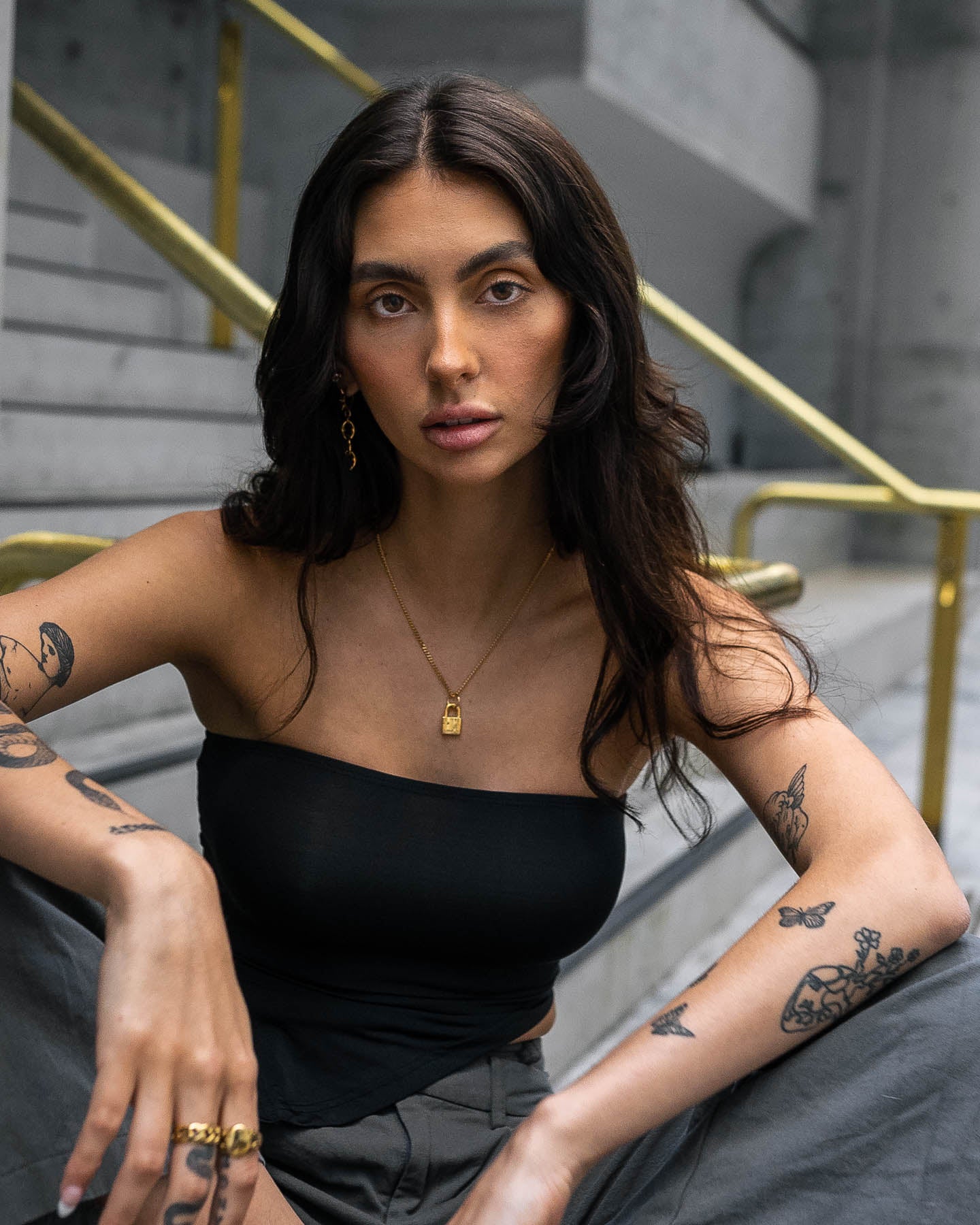 A woman with long dark hair and tattoos sits on steps with gold railings, wearing a black strapless top, gray pants, and the Mini Padlock Pendant (Gold), pairing her look with a neutral expression and elegant gold jewelry.