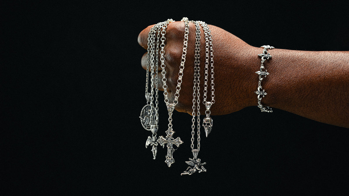 A hand holding up several silver-toned chain necklaces with various charms, including a cross, a skull, and a heart, next to a matching silver-toned bracelet, set against a solid black background.