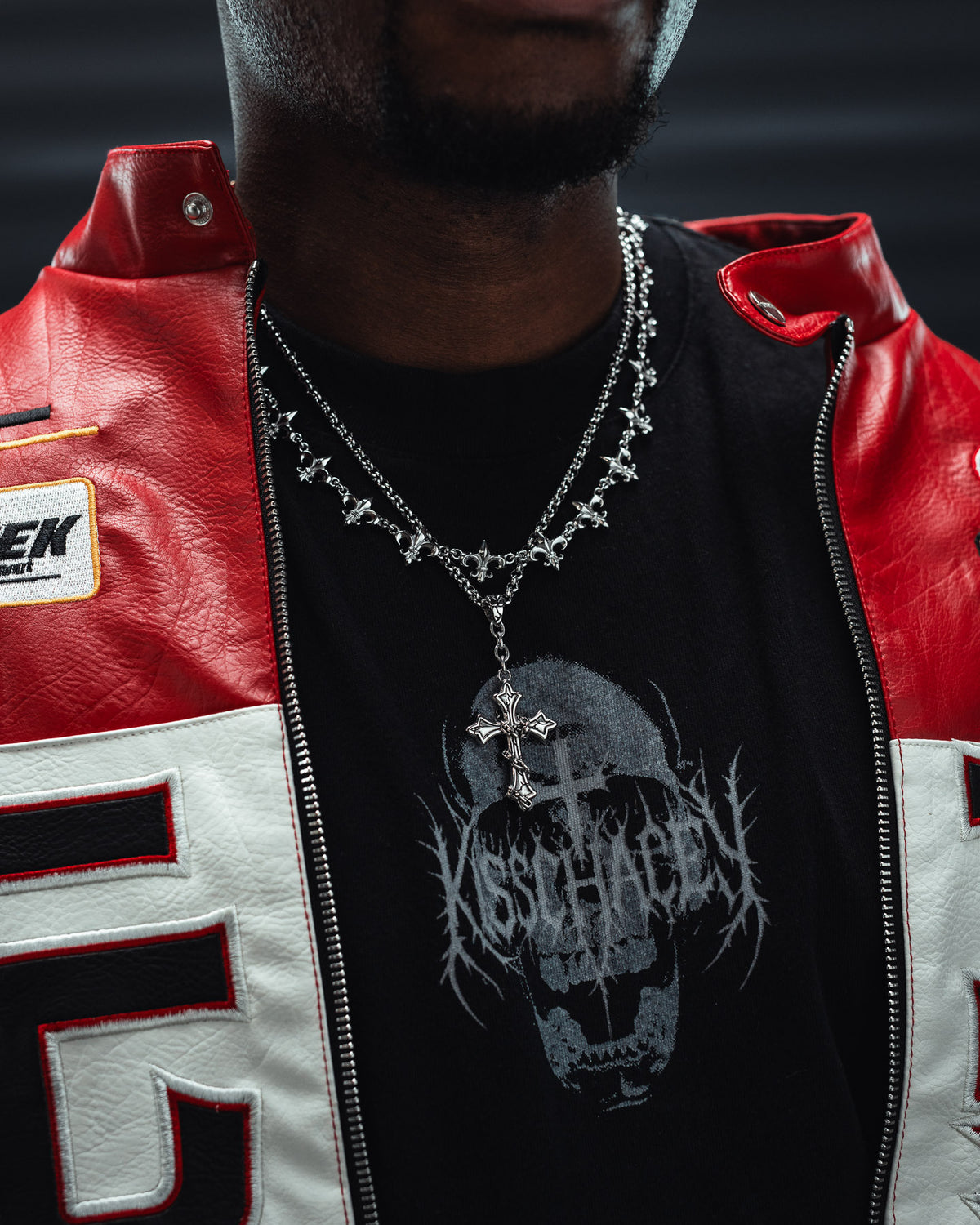 A person wearing a red and white jacket, black graphic t-shirt with a skull design, and layered silver chains featuring the Statement Collective Mini “The Ruins” Cross Pendant crafted from stainless steel jewelry. The face is partially out of frame.