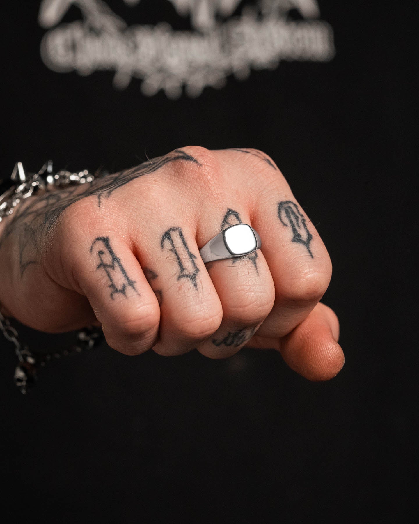 A close-up of a tattooed fist wearing the Minimal Signet Ring on the middle finger and a spiked bracelet. The dark, blurred background highlights the sleek stainless steel design.