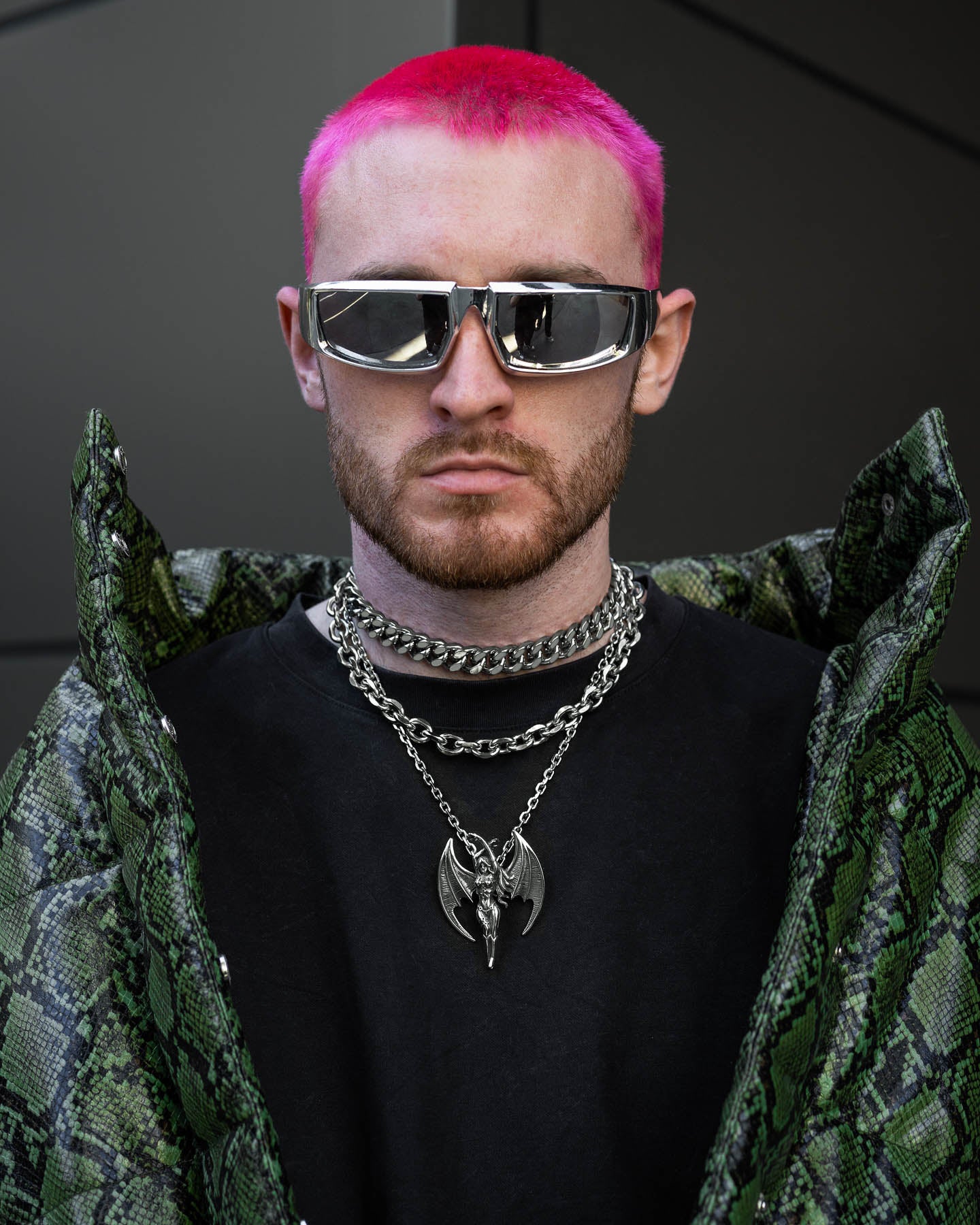A man with bright pink hair, a beard, and reflective sunglasses wears the Mistress Stack layered chain necklaces, a chunky Cuban choker, and a black shirt under a green snakeskin-patterned jacket. He stands before a dark geometric background.