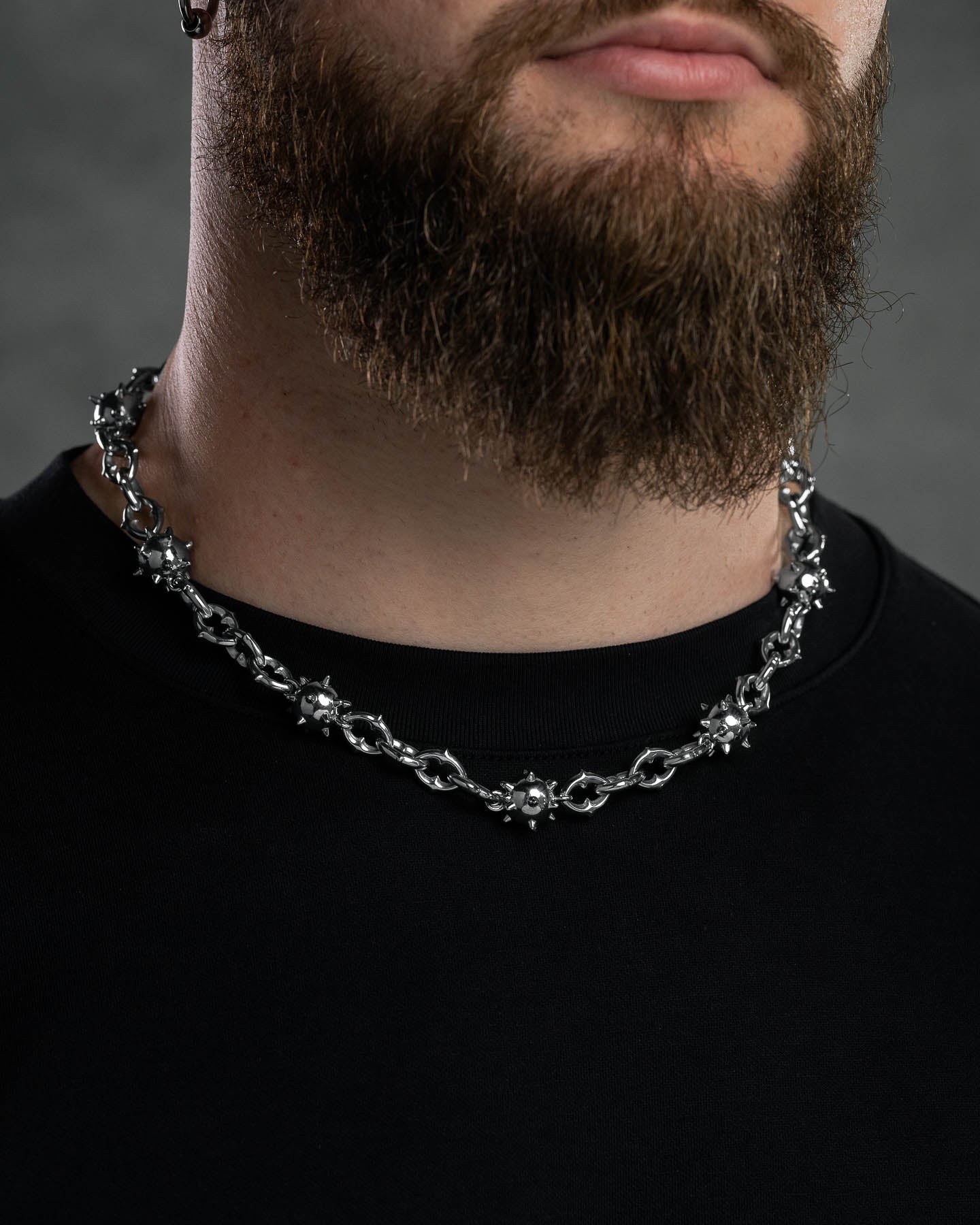 A close-up of a man with a thick brown beard wearing the "Morning Star" Cathedral Chain, a chunky stainless steel necklace with spiked ball-shaped details, and a black crew-neck shirt against an out-of-focus neutral background.