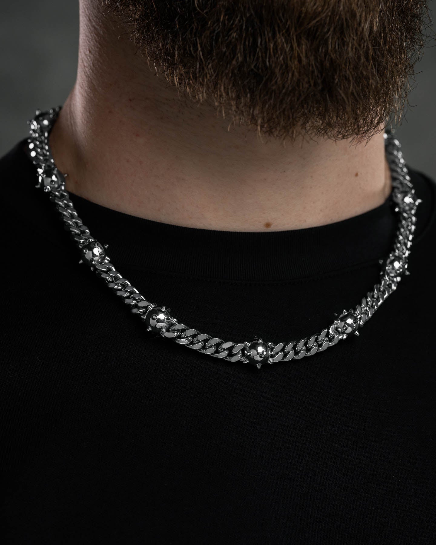 A close-up of a person wearing the "Morning Star" Cuban Chain, a thick stainless steel necklace with spike and stud details, paired with a black shirt; the lower part of their beard is visible.