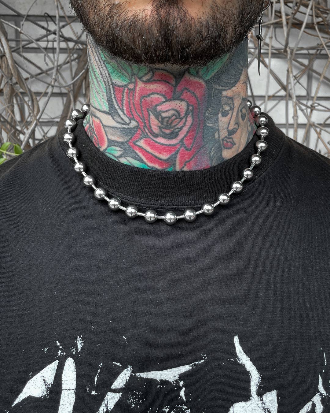 A person with colorful neck tattoos, featuring a red rose and a woman's face, wears the No Tourists Choker and a black T-shirt with white graphic text. Twigs appear in the background.