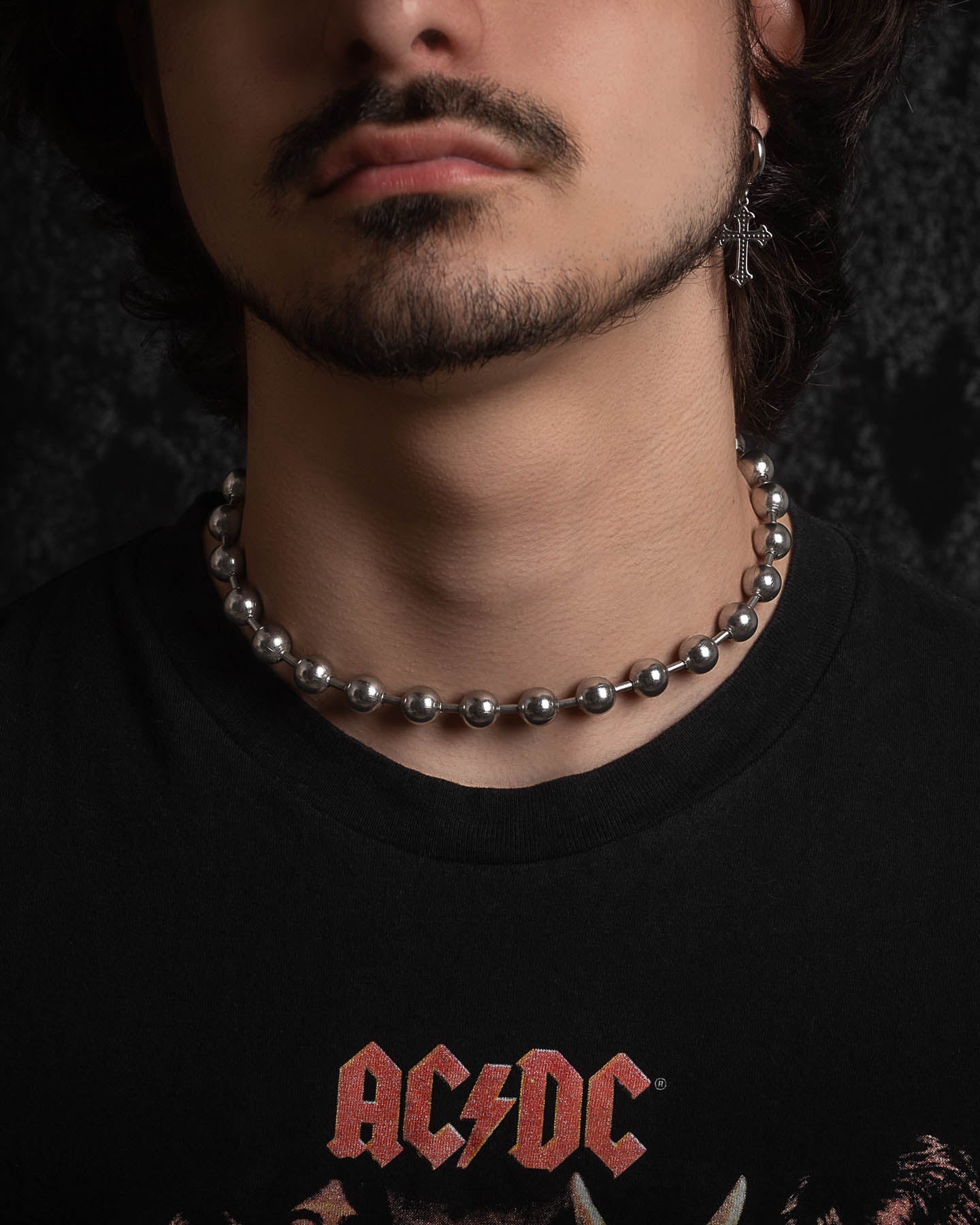 A person with a short beard and mustache wears the No Tourists Choker, a cross earring, and a black AC/DC T-shirt against a dark background.