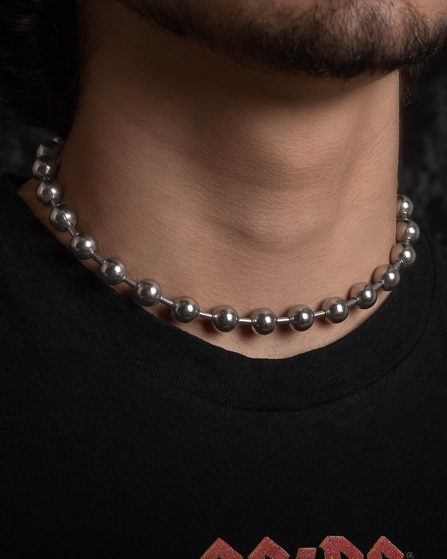 A close-up of a person's neck wearing the No Tourists Choker, a bold statement piece. The person has light facial hair and wears a black t-shirt with part of a colorful logo showing at the bottom.