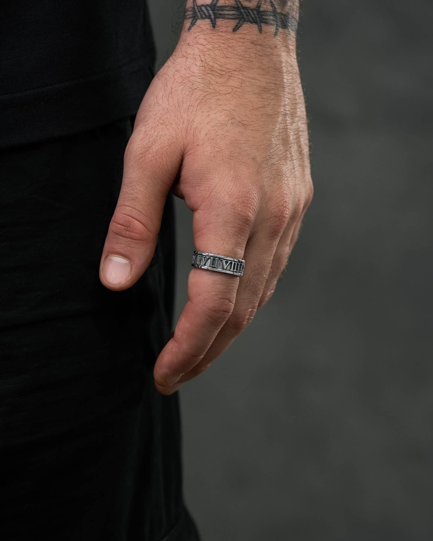 A close-up of a left hand showcases the Numerals Ring in stainless steel and a barbed wire wrist tattoo, with a dark, blurred background that highlights the sleek stacking band.