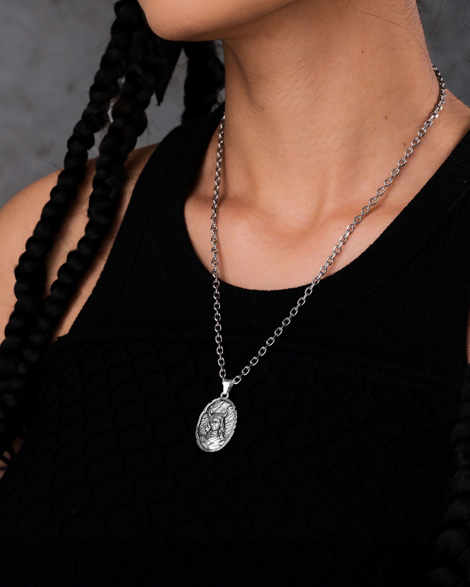 A person with long braided hair wears a sleeveless black textured top and a silver chain necklace featuring "The Unholy" Pendant, a gothic stainless steel accessory with an oval engraved design.