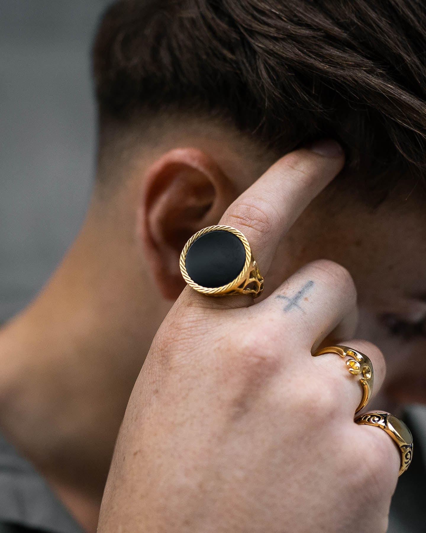 A close-up of a person touching their hair, highlighting the Onyx Sovereign Ring (Gold) with a bold black oval stone, alongside other gold rings and a small cross tattoo on their finger. The softly blurred background adds emphasis.