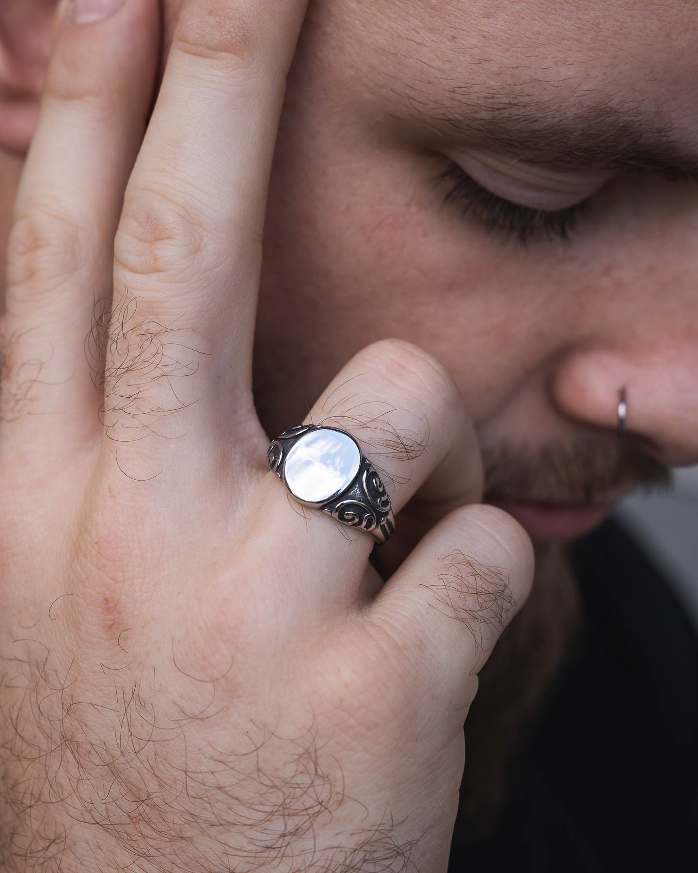 A person with a nose ring, eyes closed, touches their face while wearing the Mini Oval Scroll Signet Ring—a silver signet with a large white stone. The image highlights both the ring and their thoughtful expression.