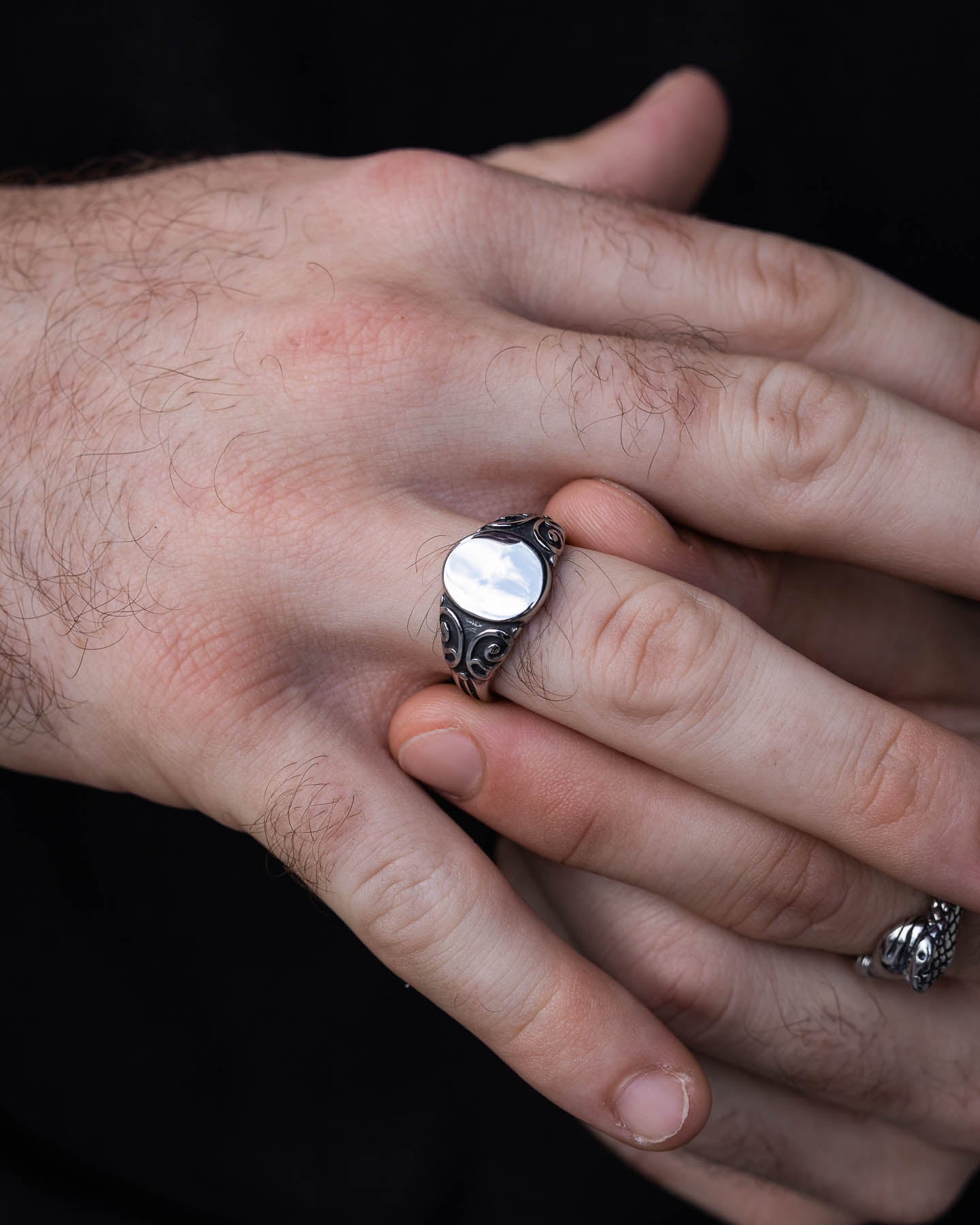 A close-up of hands, one clasping the other. The person wears the Mini Oval Scroll Signet Ring with a smooth, oval face on their index finger, along with a decorative pinkie ring on their pinky finger.