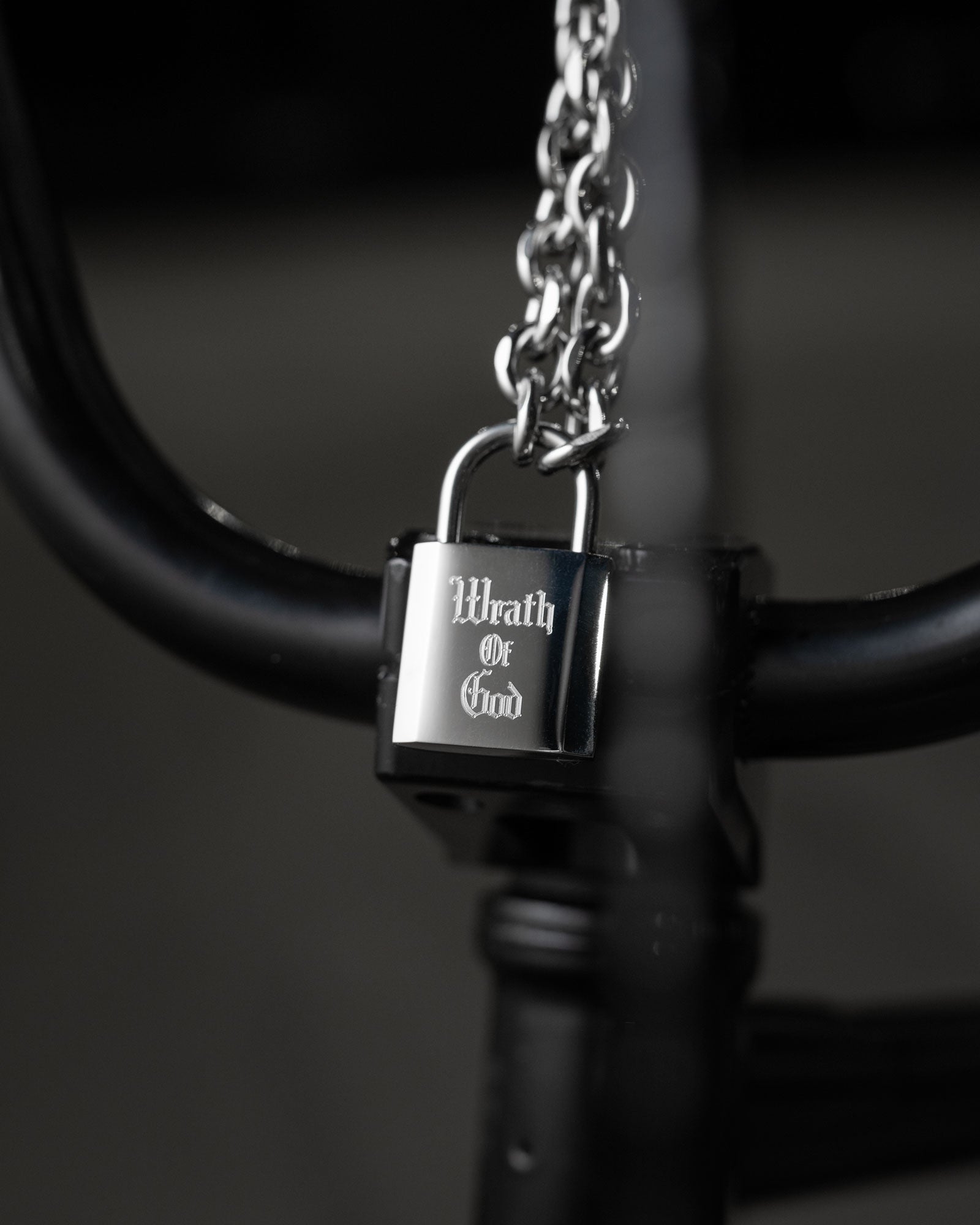 The Statement Lock & Chain 2.0, a shiny metal padlock pendant engraved with Wrath of God, hangs from a thick chain on the handlebars of a black bicycle against a blurred background.