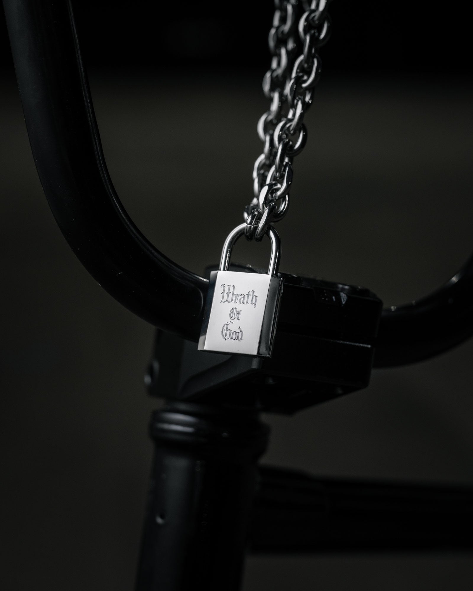 The Statement Lock & Chain 2.0, a thick metal chain with a silver padlock pendant engraved with Wrath of God, is wrapped around black bicycle handlebars against a dark, blurred background.