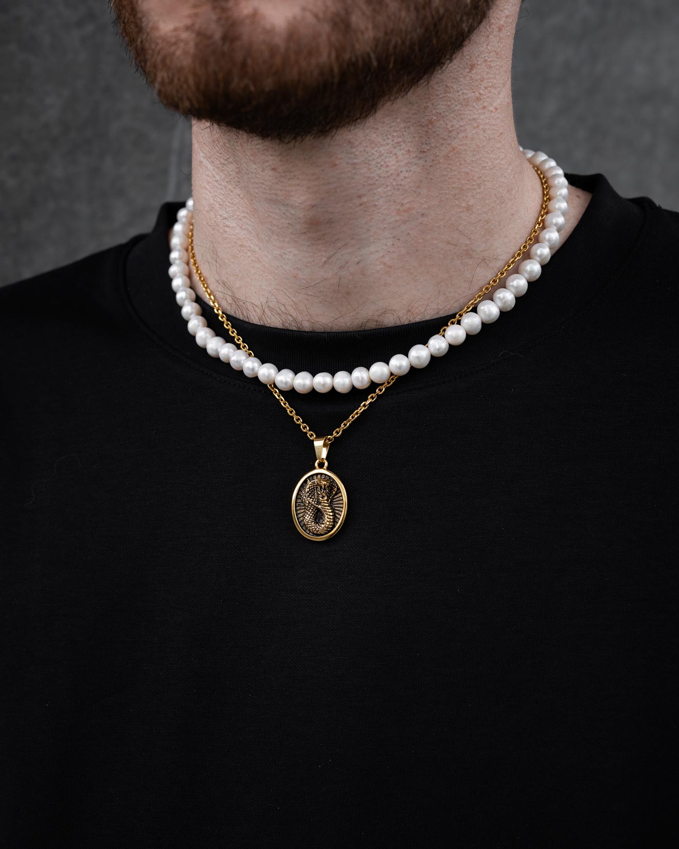 A close-up of someone wearing a black shirt and Statement Collective’s Pearl Set (Custom Pendant Choice) in gold, featuring freshwater pearls and an 18k gold pendant, set against a dark gray background.