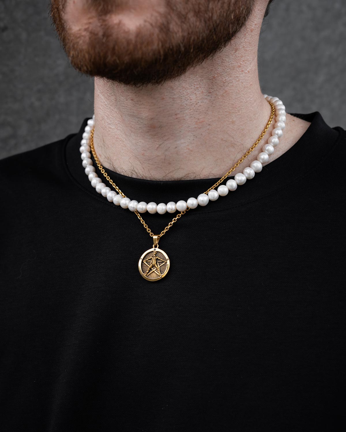 A close-up of a person wearing a black shirt and the Statement Collective Pearl Set (Custom Pendant Choice) (Gold), featuring a freshwater pearl necklace and an 18k gold pendant with a circular five-pointed star charm. Face not visible.