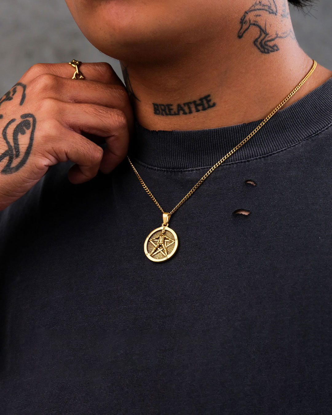 A person wearing "The Star of the Magi" Pendant (Gold) and a black shirt with small holes near the collar, showing tattoos on their hand, finger, and neck—including the word BREATHE on their neck.