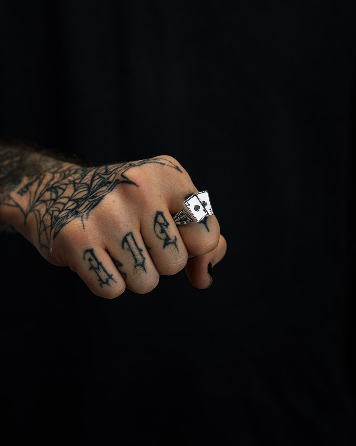 A tattooed hand with a spiderweb design and “DIE” on the fingers wears the Statement Collective Pocket Aces Ring, a stainless steel piece featuring an ace of spades motif, adding a striking touch of macabre jewellery against the dark background.
