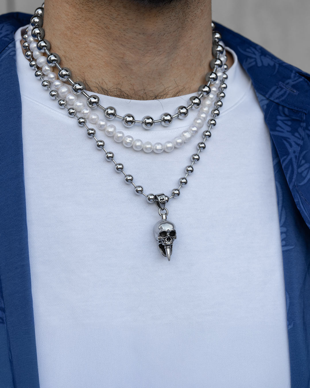 A person wears the Precious Death Stack—a bullet skull necklace with metallic beads, a pearl necklace with white beads, and a third strand of smaller metallic beads—layered over a white shirt and blue patterned jacket.